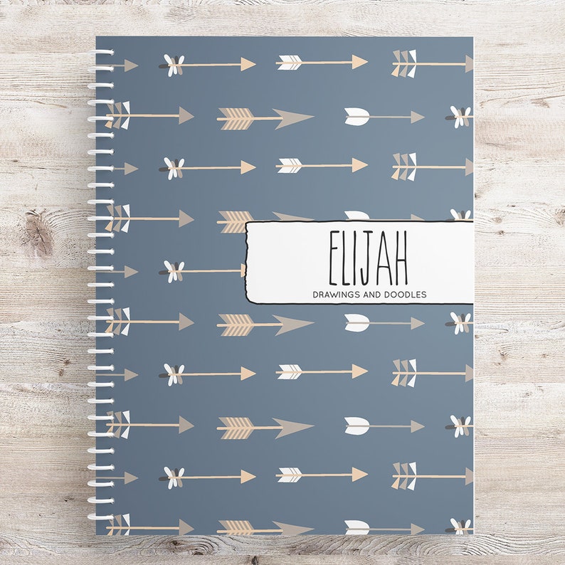 May include: A blue spiral bound notebook with a pattern of tan and white arrows. The cover has a white label with the name "ELIJAH" and the text "DRAWINGS AND DOODLES".
