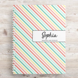 May include: A spiral bound notebook with a pink, yellow, and green striped cover. The cover has a white label with the name "Sophia" and the text "Drawings and Doodles".