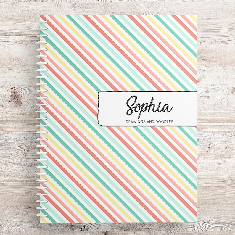May include: A spiral bound notebook with a pink, yellow, and green striped cover. The cover has a white label with the name "Sophia" and the text "Drawings and Doodles".
