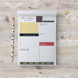 May include: A white daily planner with a black and red apple design. The planner has sections for priorities, tasks for the day, materials needed for today, schedule, and notes. The planner also has a date section with the days of the week abbreviated.