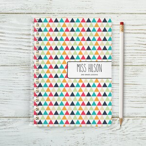 May include: A spiral bound notebook with a colourful geometric pattern of triangles in red, orange, yellow, green, blue and white. The cover has a black rectangle with the text "Miss Hilson 2nd Grade Lessons".