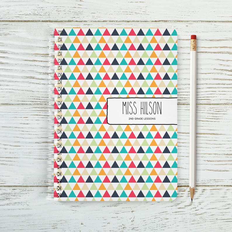 May include: A spiral bound notebook with a colourful geometric pattern of triangles in red, orange, yellow, green, blue and white. The cover has a black rectangle with the text "Miss Hilson 2nd Grade Lessons".