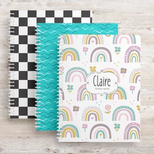 May include: Three spiral-bound notebooks with different cover designs. One has a black and white checkerboard pattern, another has teal wavy lines, and the third features a rainbow and floral design with the name "Claire" and "School Notes".