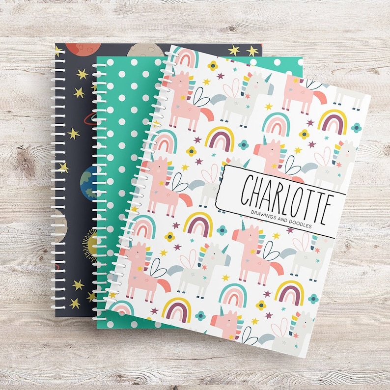May include: Three spiral bound notebooks with different covers. The front notebook has a white cover with a repeating pattern of pink and white unicorns, rainbows, and stars. The text "CHARLOTTE DRAWINGS AND DOODLES" is printed on the front cover. The middle notebook has a green cover with white polka dots. The back notebook has a dark blue cover with a repeating pattern of stars, planets, and the sun.