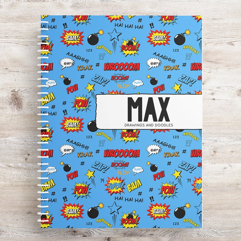 May include: A blue spiral bound notebook with a comic book theme. The cover features a repeating pattern of yellow stars, red and yellow speech bubbles with words like "BAM!" and "POW!" and black bombs. The notebook has a white label with the name "MAX" and the words "DRAWINGS AND DOODLES".