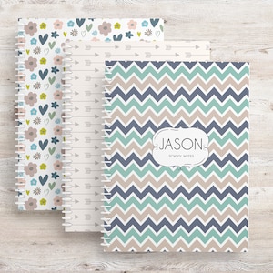 May include: Three spiral-bound notebooks with patterned covers. One features a floral design with blue, pink, and green accents. Another has a cream background with arrows. The third notebook has a blue, teal, and tan chevron pattern with the name "JASON" and "SCHOOL NOTES".