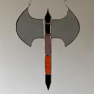 May include: A stained glass axe with a silver chain for hanging. The axe head is gray, the handle features black, pink, and orange sections. The edges are outlined in silver. A decorative piece.