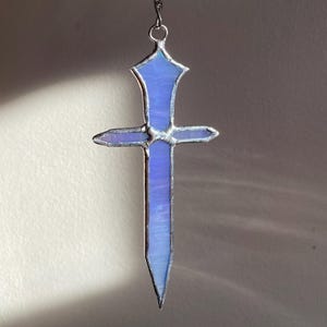 May include: A stained glass sword pendant with a light blue hue, suspended by a silver chain. The sword design features a crossguard and a pointed tip. The glass has a translucent quality, allowing light to pass through.