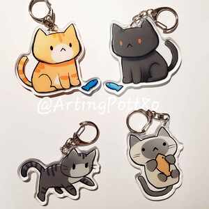 May include: Four different cat keychains with a silver key ring. The cats are in various poses and colours, including orange, black, grey, and white. The keychains are made of clear acrylic and feature a cartoon-style design. The text "@ArtingPottxo" is visible in the image.