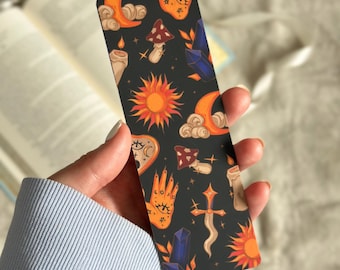 Puede incluir: Un marcapáginas negro con un patrón repetido de soles naranjas, lunas, manos, cristales y setas. El fondo es negro con estrellas blancas.