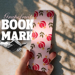 Puede incluir: Un marcapáginas rosa con un patrón de pomelo y las palabras "Grapefruit BOOK MARK". El marcapáginas presenta ilustraciones de rodajas de pomelo y hojas verdes. El marcapáginas está sostenido en una mano.