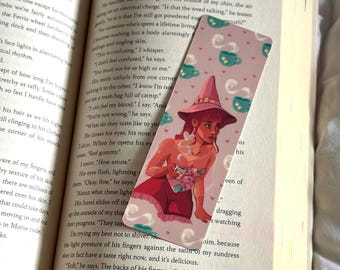 May include: A bookmark featuring a whimsical illustration of a witch holding a teacup, with a pink and white color scheme. The bookmark is inserted into a book with the title "HAPPY PLACE" visible.