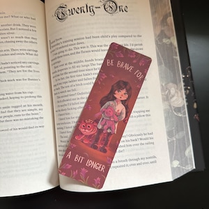 May include: A bookmark featuring the text "BE BRAVE FOR A BIT LONGER" and an illustration of a girl with a stuffed toy and a tiger. The bookmark is in an open book.