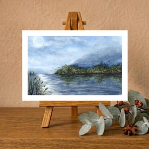 Op de afbeelding: Een aquarel op een kleine houten ezel toont een sereen meer onder een bleke maan. Het landschap bevat donkergroene bomen langs de oever, kalm blauw water dat de lucht weerspiegelt en een groep hoog riet op de voorgrond.