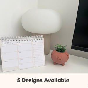 May include: A desk scene with a weekly planner labelled "THE PERFECT WEEK" and a pig-shaped plant pot. A white lamp and the text "5 Designs Available" are also visible. The planner is spiral-bound.
