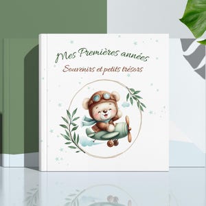 Peut inclure: Un livre de souvenirs pour bébé à couverture blanche avec le texte français "Mes Premières années, Souvenirs et petits trésors". La couverture présente une illustration d'ours en peluche pilote sur un biplan, entouré d'une couronne.