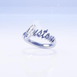 May include: A silver ring with the word "Custom" in a cursive font. The ring is displayed on a white surface with a soft blue gradient background. The ring is a piece of jewelry.
