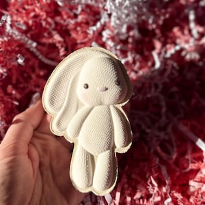 May include: A white bunny-shaped decorative item with a textured surface. The bunny has large, floppy ears, a small nose, and tiny brown eyes. The item is held in a hand against a background of red and white shredded paper.
