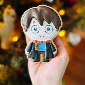 May include: A Harry Potter-themed cookie, featuring a character design with brown hair, glasses, and a lightning bolt scar. The cookie has a black robe, a blue shirt, and a scarf with red and gold stripes. The cookie is held in a hand.