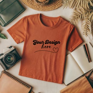 Könnte beinhalten: Ein orangefarbenes T-Shirt mit kurzen Ärmeln und einem Rundhalsausschnitt. Das Shirt hat den Text "Your Design here" in Schwarz mit einem kleinen Herz auf der Vorderseite.