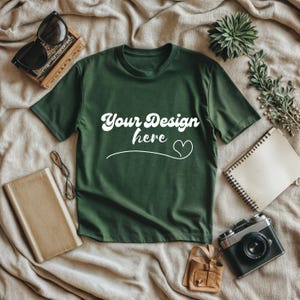 Op de afbeelding: Donkergroen t-shirt met de tekst "Your Design here" in witte letters. Het shirt is omgeven door accessoires, waaronder een zonnebril, een notitieboek, een camera en een kleine leren tas.