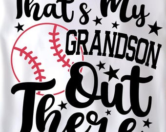 May include: White t-shirt with the text "That's My Grandson Out There" in black script, with a baseball graphic in red and white. The shirt is folded and displayed on a wooden plate, with greenery and red berries around the plate.
