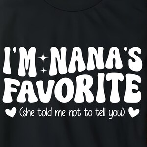 May include: Black t-shirt with the text "I'M NANA'S FAVORITE" in white, retro-style font. Below the text is "(She told me not to tell you)" in a smaller font. Two white hearts flank the text. The shirt is on a wooden tray.