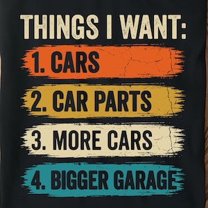 Puede incluir: Camiseta negra con el texto "THINGS I WANT: 1. CARS, 2. CAR PARTS, 3. MORE CARS, 4. BIGGER GARAGE" en una fuente desgastada. El texto está superpuesto a pinceladas horizontales coloridas. La camiseta se muestra en una bandeja de madera.