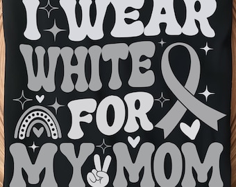 Puede incluir: Camiseta negra con el texto "I Wear White For My Mom" en blanco y gris. El diseño incluye una cinta gris, un arcoíris, corazones y un signo de la paz. La camiseta se muestra en una bandeja de madera.