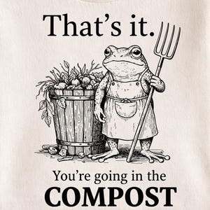 May include: Cream-coloured t-shirt with a black and white illustration of a frog holding a pitchfork. The frog stands beside a wooden barrel filled with plants. Text on the shirt reads "That's it. You're going in the COMPOST."