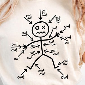 May include: Cream-colored t-shirt featuring a stick figure with an 'X' for eyes and a distressed expression. Black arrows point towards the figure, each accompanied by the word 'ow!' in a playful, cartoonish style. The shirt's design is simple and humorous.