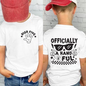 May include: White t-shirts with black graphics. One shirt features a hand graphic with the text "HIGH FIVE". The other shirt has the text "OFFICIALLY A HAND FUL" with sunglasses, thumbs up, and stars. The number 5 is also included.