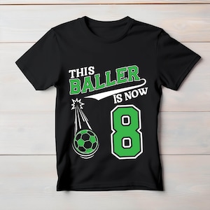 Puede incluir: Camiseta negra con el texto "THIS BALLER IS NOW 8" en blanco y verde. Un gráfico de un balón de fútbol con un diseño verde y negro se muestra con un efecto de estrella fugaz. El número 8 es verde.
