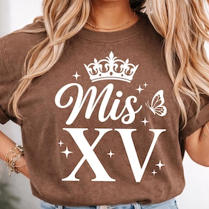 May include: Brown t-shirt with white text and graphics. The design includes the words "Mis XV" with a crown, butterfly, and star accents. The shirt has rolled-up sleeves and a crew neck.