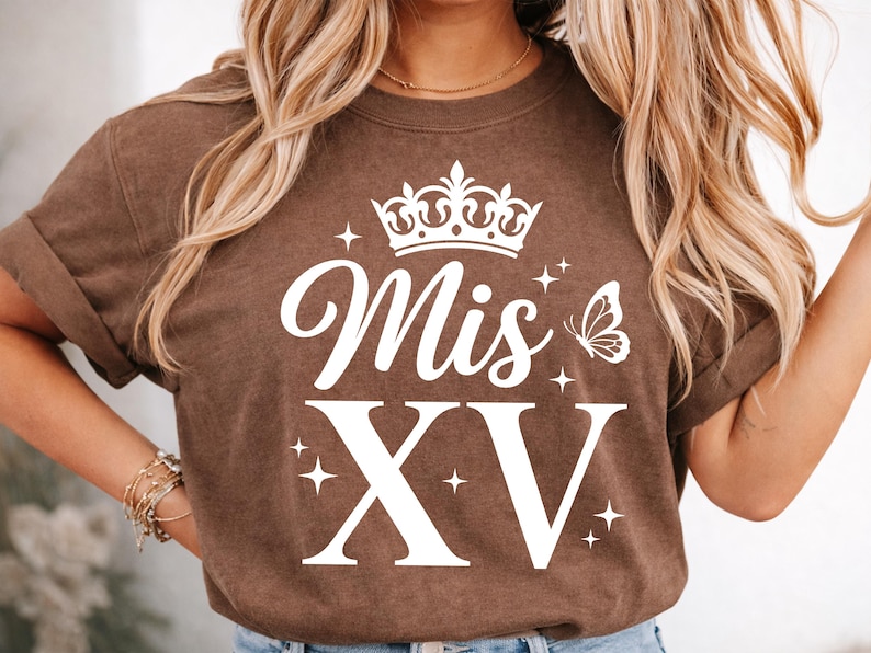 May include: Brown t-shirt with white text and graphics. The design includes the words "Mis XV" with a crown, butterfly, and star accents. The shirt has rolled-up sleeves and a crew neck.