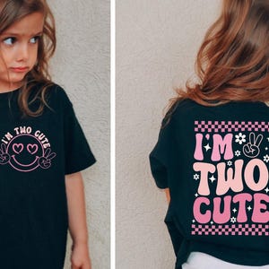 Puede incluir: Camiseta negra con el texto "I'M TWO CUTE" en rosa y blanco. La parte delantera presenta una carita sonriente con ojos de corazón y signos de paz. La parte trasera tiene el mismo texto con un patrón de tablero de ajedrez y flores.