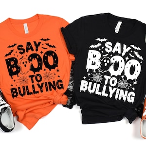 May include: Two Halloween-themed t-shirts, one orange and one black, with the phrase "Say Boo to Bullying" in a spooky font. The design includes bats, a ghost, and spiderwebs. Black and white sneakers and orange sneakers are visible.