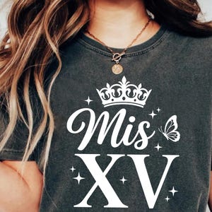 May include: Dark gray t-shirt with a white crown, the words "Mis XV", a butterfly, and stars. The shirt has a round neckline and short sleeves. The text is in a script font.