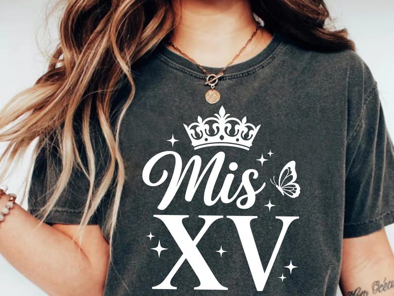May include: Dark gray t-shirt with a white crown, the words "Mis XV", a butterfly, and stars. The shirt has a round neckline and short sleeves. The text is in a script font.