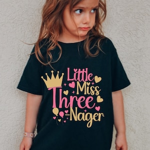 May include: Black t-shirt with the text "Little Miss Three Nager" in pink and yellow. The design includes a gold crown, hearts, and balloons. The shirt is worn by a young child.