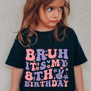 Op de afbeelding: Zwart T-shirt met de tekst "BRUH IT'S MY 8TH BIRTHDAY" in roze en paarse retro letters. Het ontwerp bevat harten, bloemen en een vredesteken. Het T-shirt wordt door een kind gedragen.