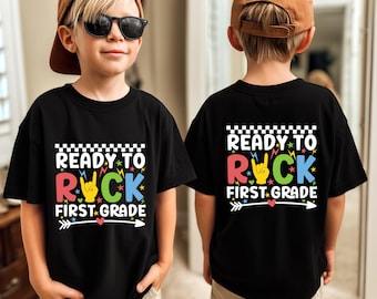 May include: Black t-shirt with the text "READY TO ROCK FIRST GRADE" in a colorful design. The design includes a rock and roll hand gesture, stars, and an arrow. The shirt is worn by a child wearing sunglasses and a brown cap.