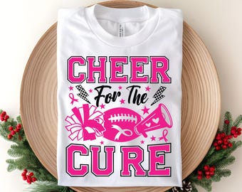 Op de afbeelding: Wit T-shirt met een roze en zwart ontwerp met de tekst "CHEER FOR THE CURE". Het ontwerp bevat een voetbal, megafoon, pompon, bliksemschichten en roze linten. Het shirt wordt gepresenteerd op een houten dienblad.