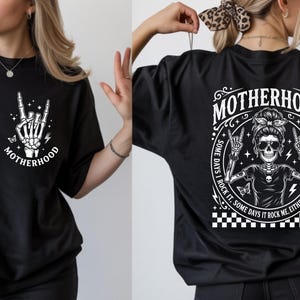 May include: Black t-shirt featuring a white graphic of a skeleton hand making a rock on gesture, with the word "MOTHERHOOD". The back of the shirt has a circular design with a skeleton wearing sunglasses and the text "MOTHERHOOD".