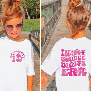 May include: White t-shirt with pink graphics. The front features the text "IN MY DOUBLE DIGITS ERA" with a crown, heart, and the number 10. The back of the shirt has the same text with a butterfly and flower design.