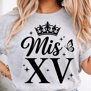 May include: A light gray t-shirt with the words "Mis XV" in black script and block letters, topped with a black crown and a butterfly. The shirt also features small star accents.