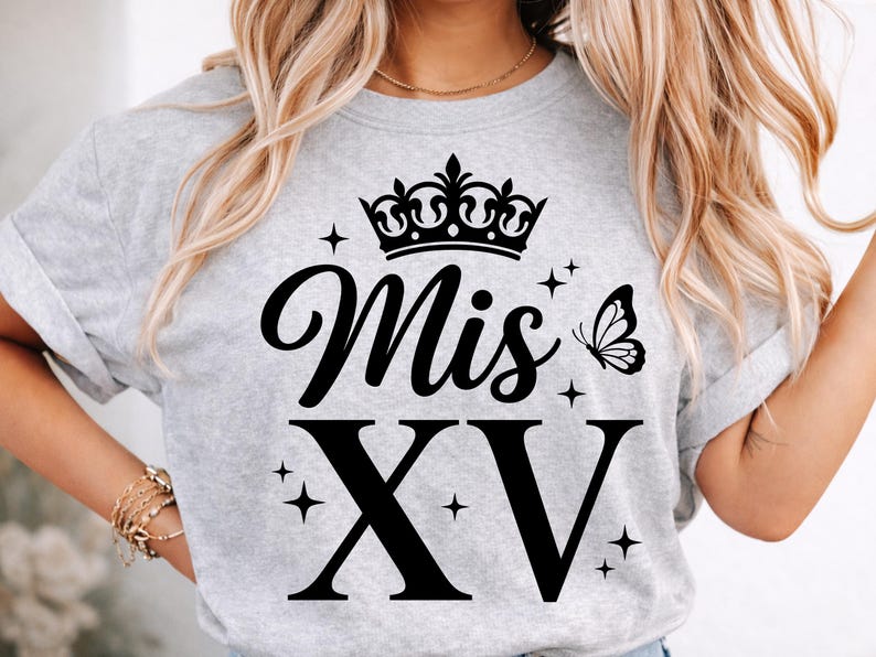 May include: A light gray t-shirt with the words "Mis XV" in black script and block letters, topped with a black crown and a butterfly. The shirt also features small star accents.