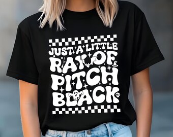 Puede incluir: Camiseta negra con la frase "Just a Little Ray of Pitch Black" en una fuente retro. El diseño incluye estrellas, un murciélago, un sombrero de bruja, un fantasma y una calavera. La parte superior e inferior de la camiseta tienen un patrón de cuadros.