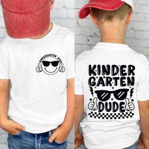 Puede incluir: Camiseta blanca con un gráfico negro en la parte delantera y trasera. La parte delantera presenta una carita sonriente con gafas de sol con el texto "Kindergarten Dude". La parte trasera tiene las palabras "Kindergarten Dude" con gafas de sol, rayos y pulgares arriba.