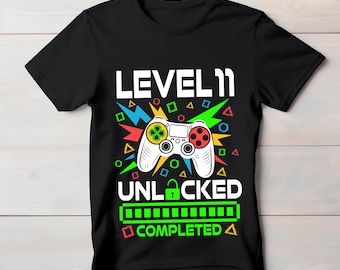 Könnte beinhalten: Schwarzes T-Shirt mit einem weißen Aufdruck, der einen Videospiel-Controller und den Text "LEVEL 11 UNLOCKED COMPLETED" zeigt. Der Controller hat grüne, gelbe und rote Knöpfe. Bunte geometrische Formen umgeben das Design.